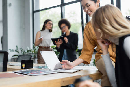 smiling asian businesswoman looking at laptop near blurred colleague in officeの写真素材