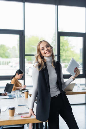 cheerful businesswoman with digital tablet looking at camera near interracial colleagues working on blurred backgroundの写真素材