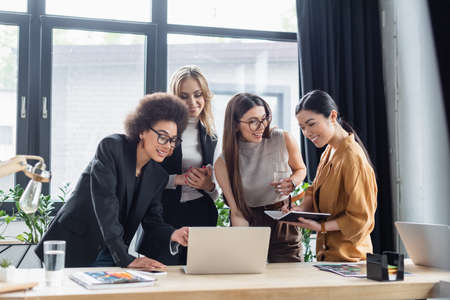 cheerful african american businesswoman pointing at laptop near multicultural coworkers