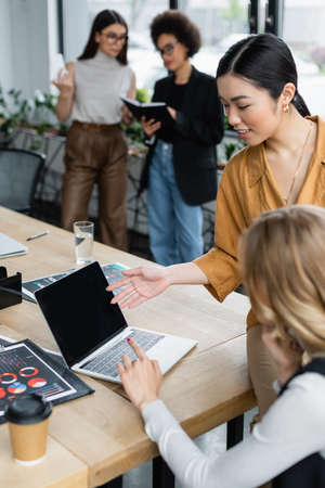 asian businesswoman pointing at laptop near blurred manager and infographics