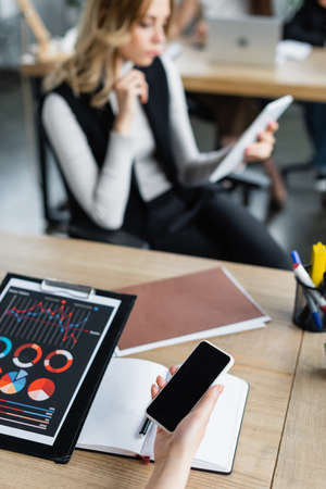 blurred businesswoman using digital tablet near colleague with smartphone and chartsの写真素材
