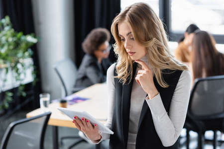 thoughtful businesswoman looking at digital tablet near multiethnic colleagues working on blurred...