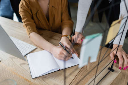 partial view of businesswoman writing in empty notebook near colleague at workplaceの写真素材