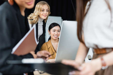 interracial businesswomen working near computer monitor and colleagues on blurred foregroundの写真素材