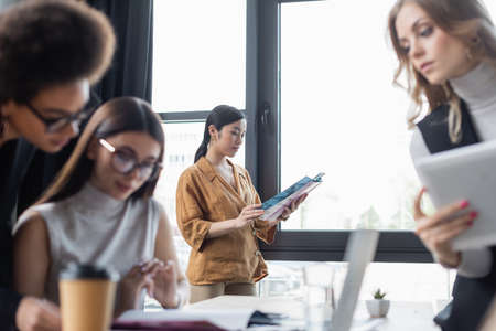 young asian businesswoman reading magazine while colleagues working on blurred foregroundの写真素材