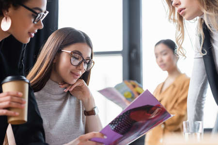 brunette businesswoman smiling while reading magazine near multiethnic coworkersの写真素材