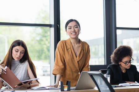 happy asian manager smiling at camera near interracial colleagues at workplaceの写真素材