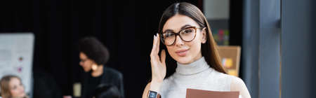 brunette businesswoman touching eyeglasses while looking at camera in office, banner