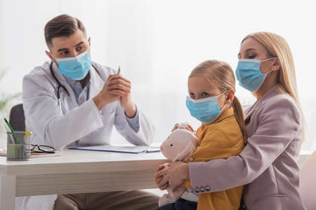 Kid looking away near mother in medical mask and family doctor in clinicの写真素材