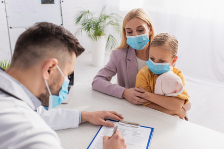 Woman hugging child with toy near pediatrician writing on clipboard in hospitalの写真素材