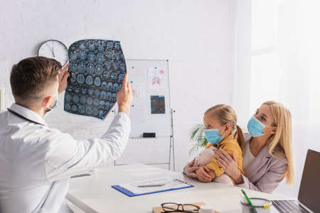 Pediatrician holding mri scan near mother and daughter in medical masks in clinic