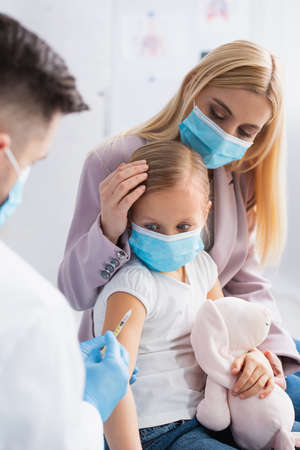 Blurred pediatrician holding syringe near mother and girl in medical masksの写真素材