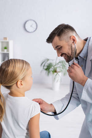 Cheerful pediatrician examining kid with stethoscope in hospitalの写真素材
