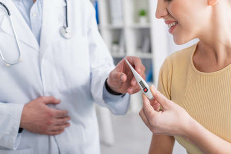 partial view of smiling woman taking electronic thermometer from doctor in clinic