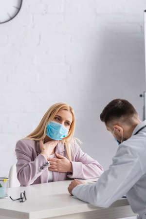 diseased woman in medical mask touching chest and throat near doctorの写真素材