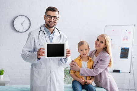 Positive family doctor holding digital tablet near mother embracing girl on medical couchの写真素材