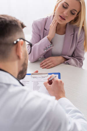 blurred doctor writing prescription near blonde woman in hospital