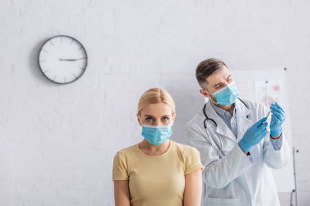blonde woman in safety mask looking at camera near physician filling syringe with vaccine