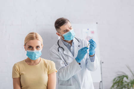 doctor in latex gloves holding vaccine near woman in medical mask in hospital