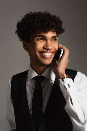 trendy african american man smiling while talking on mobile phone isolated on gray