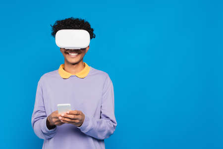 cheerful african american man using smartphone in vr headset isolated on blue