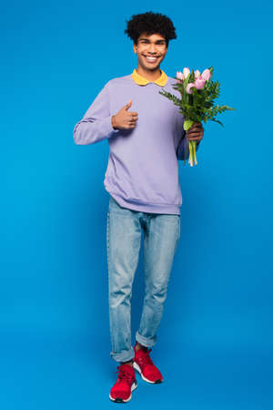 pleased african american man with bouquet of tulips showing thumb up on blue backgroundの写真素材