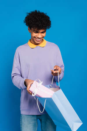 cheerful african american man putting present into paper bag isolated on blueの写真素材