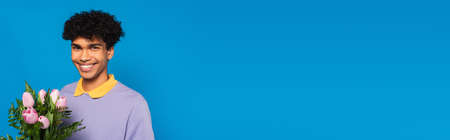 happy african american man with bouquet of pink tulips and green leaves isolated on blue, bannerの写真素材