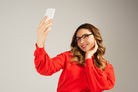 joyful woman in eyeglasses taking selfie on smartphone isolated on grayの写真素材