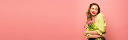 cheerful curly woman holding bouquet of flowers isolated on pink, banner