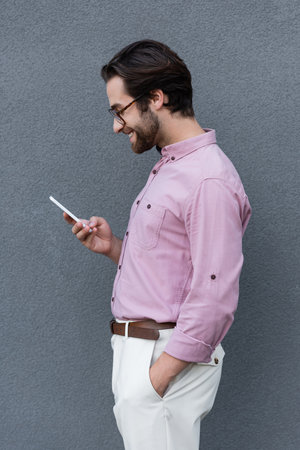Side view of smiling businessman using smartphone near wall outdoorsの写真素材
