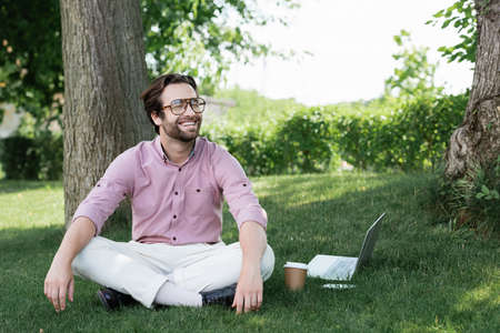 Happy businessman sitting near devices and paper cup on grass