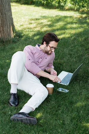 High angle view of smiling businessman using laptop near takeaway drink and smartphone on grass