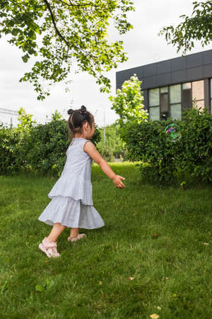 asian girl in dress walking on grass and looking at soap bubble outsideの写真素材
