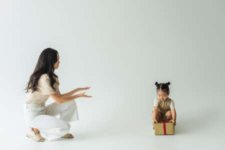 asian toddler girl holding present near brunette mother with outstretched hands on grayの写真素材