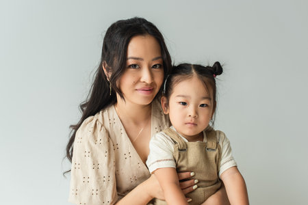 cheerful asian woman hugging toddler daughter isolated on grayの写真素材