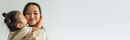 asian toddler girl kissing cheek of happy mother isolated on gray, banner