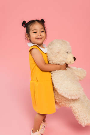 happy asian girl in yellow dress holding teddy bear on pinkの写真素材