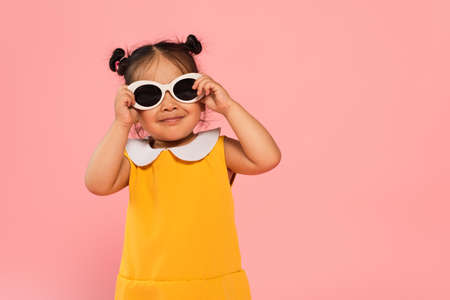 cheerful asian toddler kid in yellow dress adjusting sunglasses isolated on pinkの写真素材