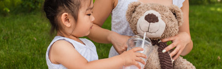 asian toddler girl holding plastic cup with milkshake near teddy bear in hands of mother, bannerの写真素材