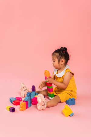 brunette asian toddler girl in dress playing building blocks on pinkの写真素材