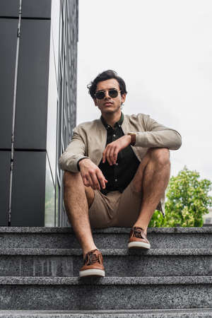 full length of young man in stylish sunglasses and bomber jacket sitting on stairs near buildingの写真素材