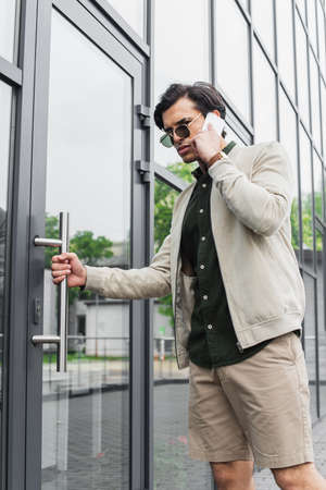 stylish young man in sunglasses talking on mobile phone while entering building