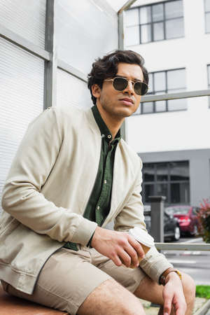young man in stylish sunglasses and bomber jacket holding coffee to go outside