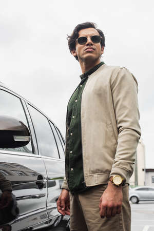 low angle view of young man in stylish sunglasses and bomber jacket posing near modern car outside