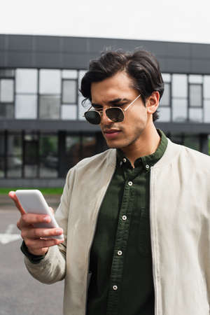 stylish young man in sunglasses and bomber jacket texting on smartphone near building