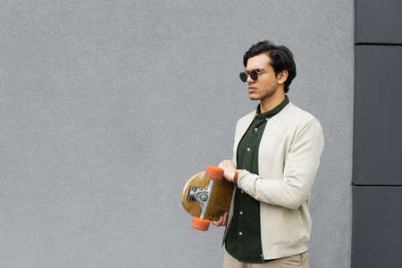 trendy young man in sunglasses holding longboard near building