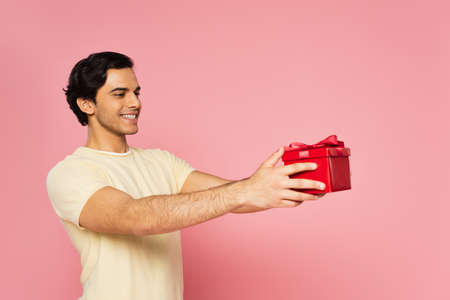 young joyful man holding red gift box isolated on pinkの写真素材