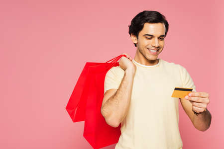 happy young man holding red shopping bags while looking at credit card isolated on pinkの写真素材