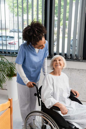 African american nurse looking at elderly patient in gown and wheelchair